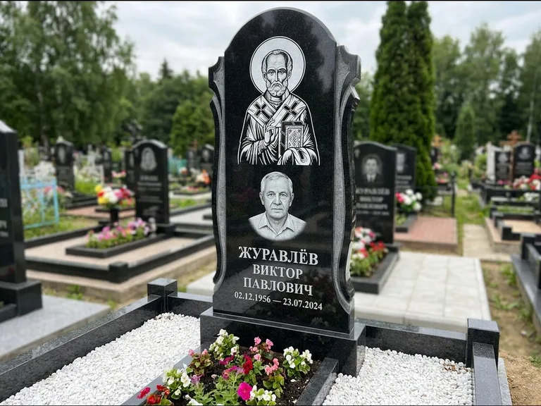 Памятник с гравировкой иконы: стела с образом в полный рост на обороте — гравировка образа Матроны Московской, низкие кусты спиреи в цвету, работа «Лебедева» в Ульяновске